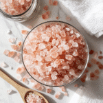 Beautiful overhead flat lay of pink Himalayan salt crystals, glass jar with sole water, wooden spoon, and essential oils on white marble surface for pink salt trick wellness guide